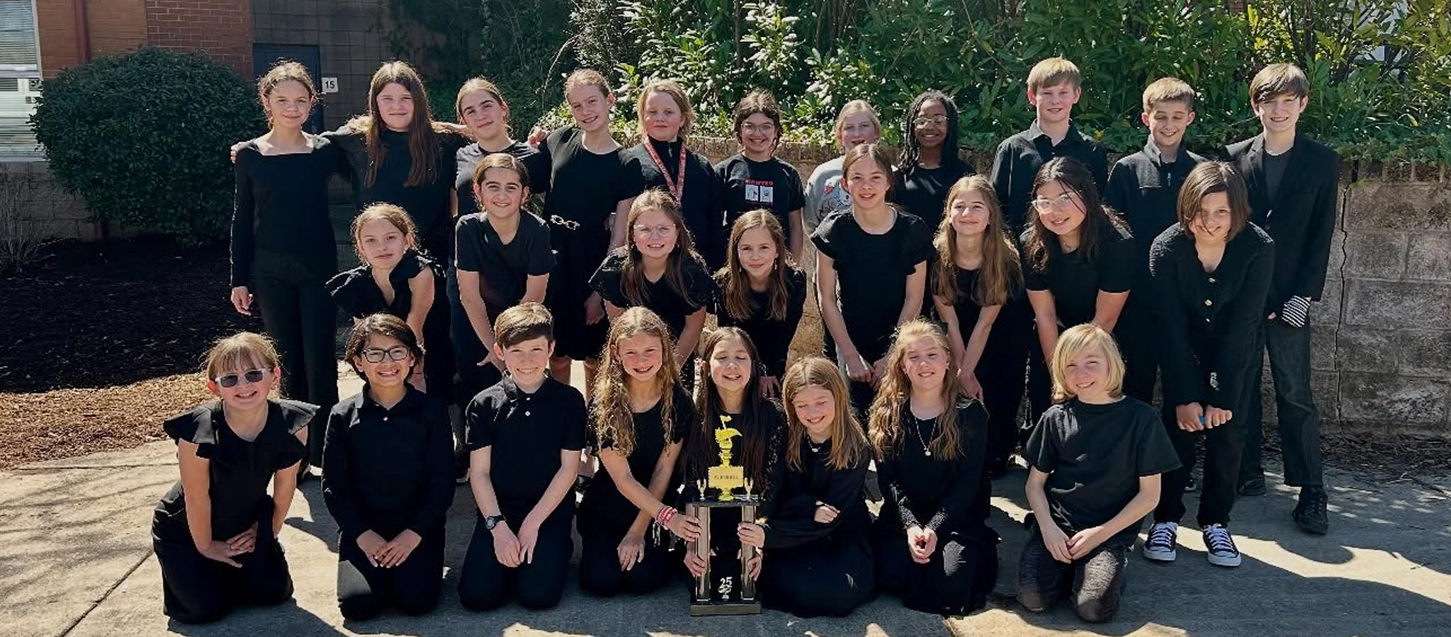 Large group of students in all black in front of trophy