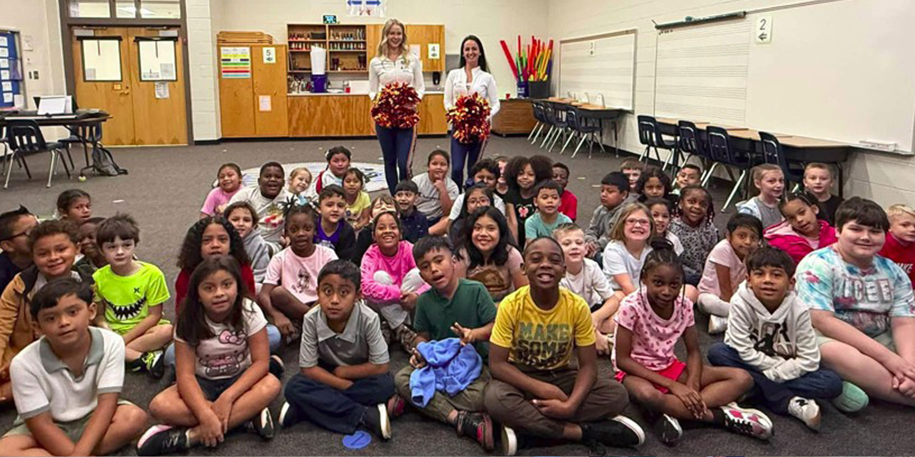 group of students sitting in their classroom with two teachers in the back holding red pom poms